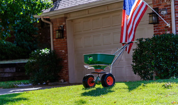 A green and white fertilizer spreader in front of a house with an American flag flying